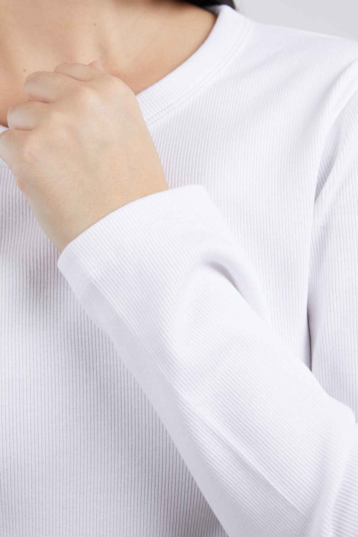 A close-up of a person wearing the Foxwood Scoop Long Sleeve Rib shirt in white. The person is partially visible, with their hand touching the Foxwood Scoop neckline of the shirt. The focus is primarily on the rib material's texture and color on the sleeve and upper chest area.