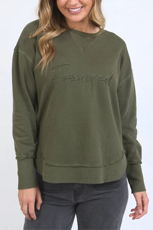 A woman wears the Foxwood Signature Crew, an olive green 100% cotton crewneck with "Foxwood" embroidered on the front, paired with dark gray jeans. Her wavy light brown hair frames her cheerful smile.