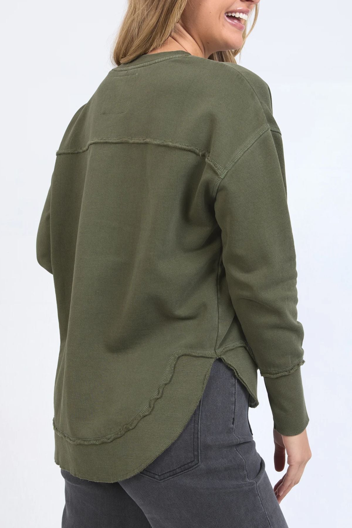 A woman in a Foxwood Signature Crew, an olive green 100% cotton long-sleeve top with raw hem details, pairs it with dark gray jeans and smiles while facing away from the camera.