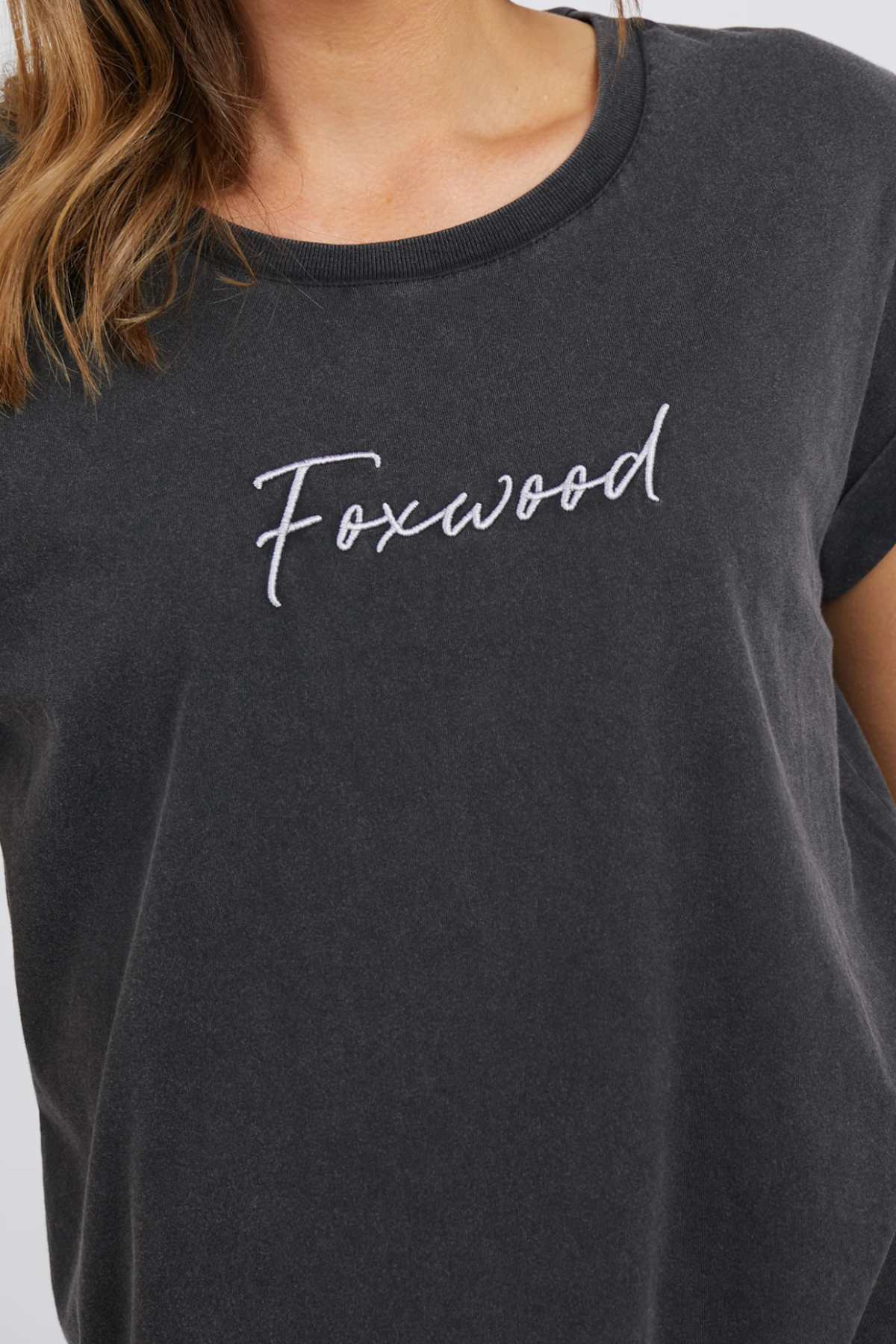 A close-up of a person wearing the Foxwood Signature Tee, a dark gray T-shirt with the word "Foxwood" embroidered in white cursive letters on the chest. Only part of the person's head and shoulder is visible.