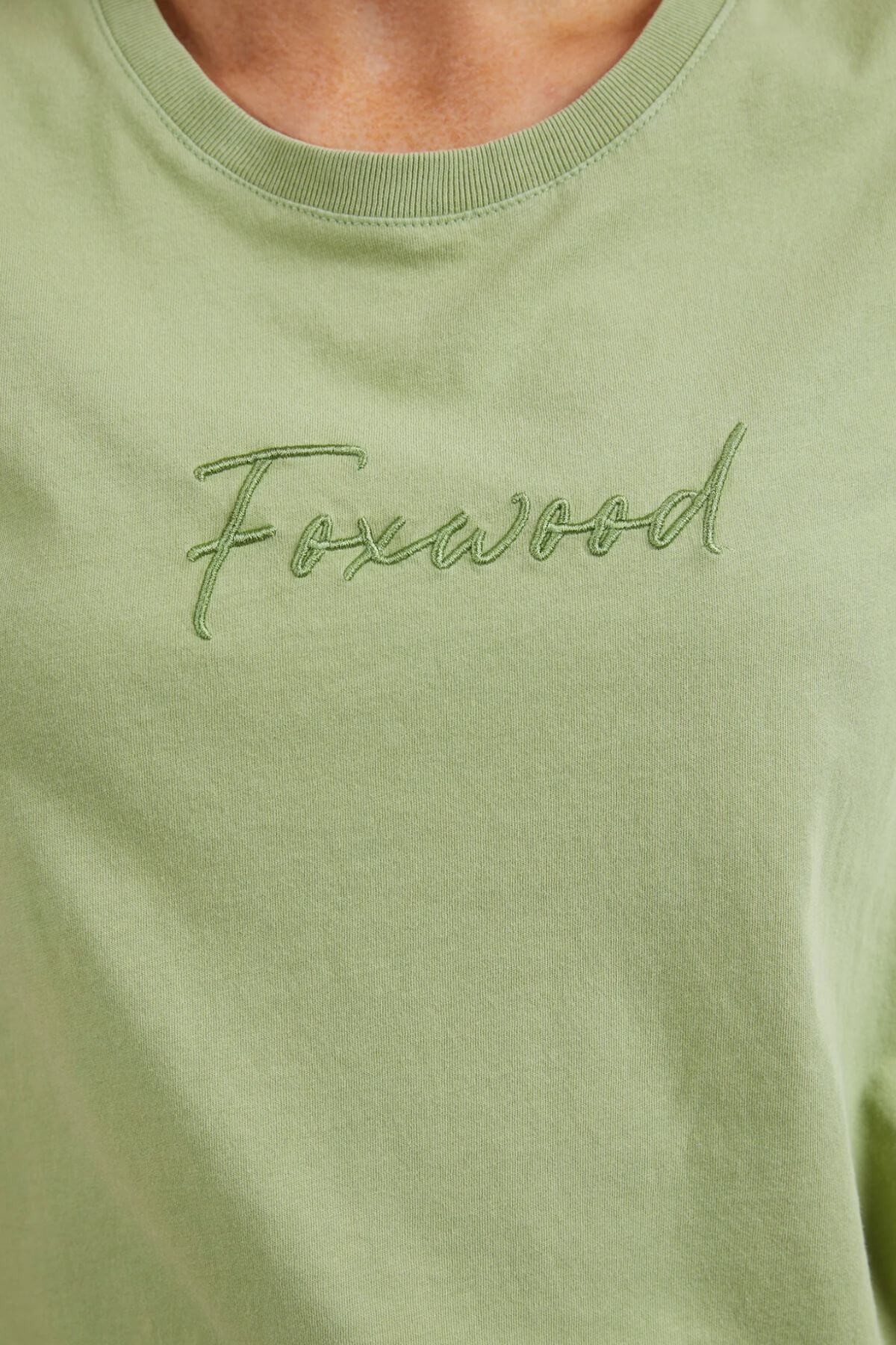 Close-up of the Foxwood Signature Tee, featuring "Foxwood" embroidered in matching thread on a light green 100% cotton T-shirt. The breathable material showcases fabric texture and fine stitching.