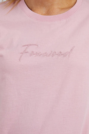 Close-up of the Foxwood Signature Tee, a light pink casual top featuring "Foxwood" embroidered in cursive on the chest. Crafted from 100% cotton for all-day comfort.