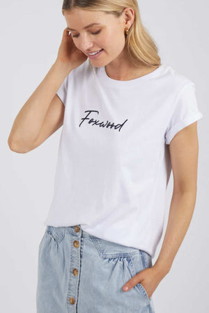 A person with long blonde hair is wearing a Foxwood Signature Tee, which is a white t-shirt with "Forward" written on it in black cursive text. They are also wearing light blue jeans with button details. The individual is smiling and has their right hand touching their head.