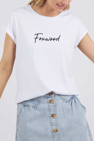 A person wearing the Foxwood Signature Tee, a white T-shirt with the word "Foxwood" printed in black cursive font in the center. The individual is also sporting a light blue, high-waisted, button-down denim skirt. The image shows the upper half of the person's body.