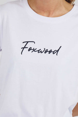 A person wearing the Foxwood Signature Tee, a white T-shirt with "Foxwood" printed in a cursive font on the front. The image focuses on the upper torso, showcasing the text prominently.