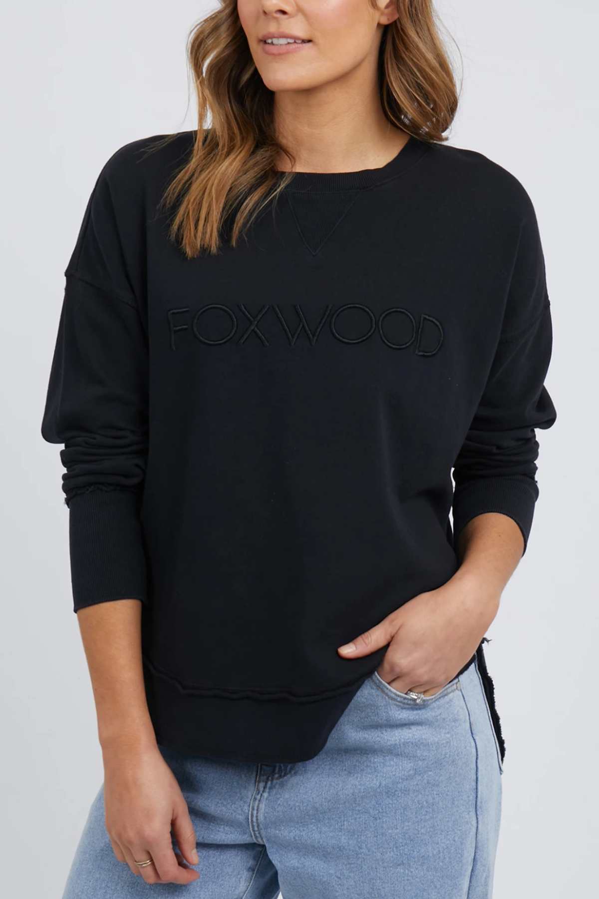 A woman with wavy, shoulder-length hair is seen wearing a black Foxwood Simplified Crew sweatshirt, elegantly embroidered with "FOXWOOD" on the front. She complements it with light blue jeans and has one hand casually placed in her pocket against a plain white background.