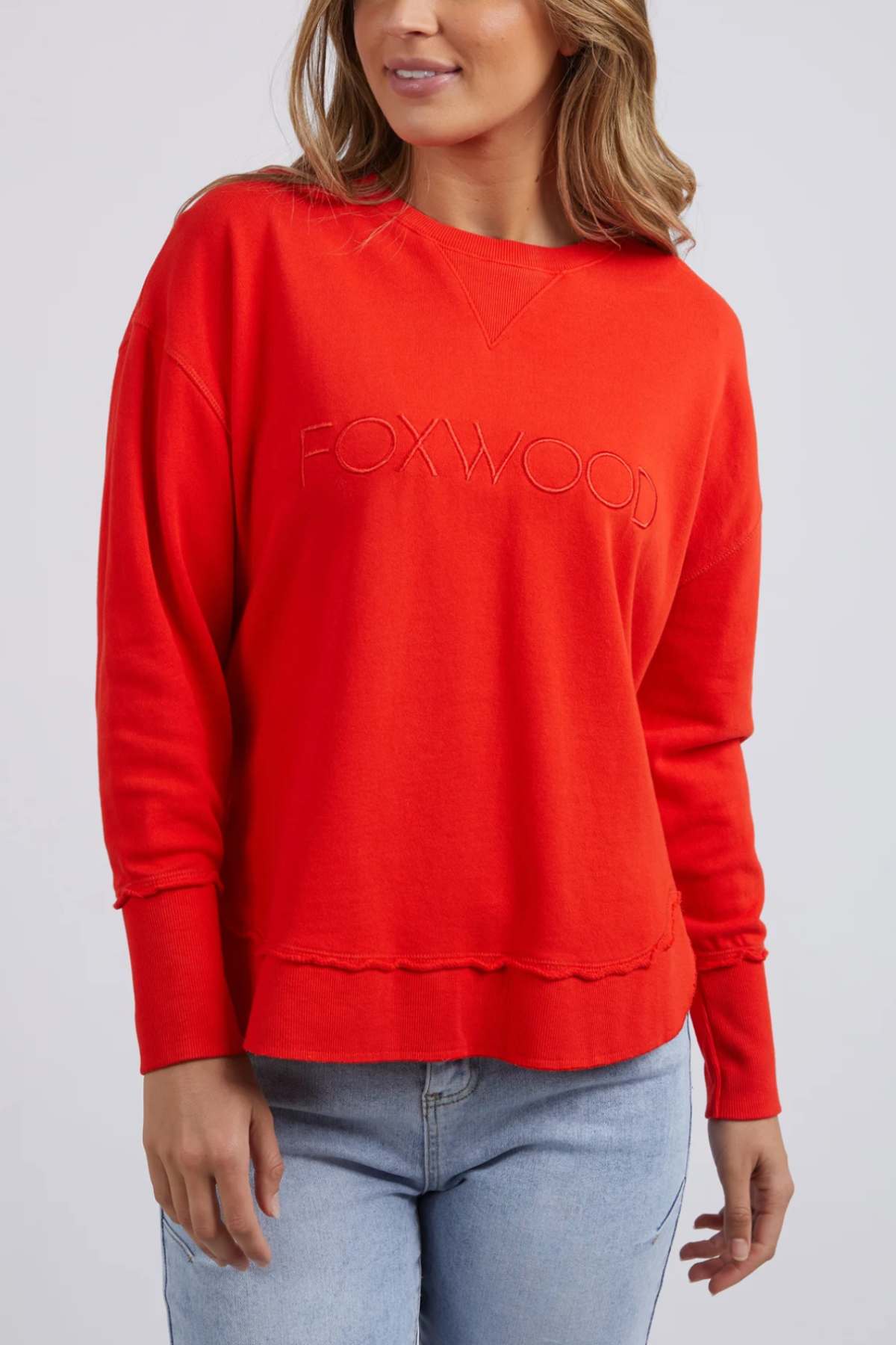 A person in a vivid red Foxwood Simplified Crew sweatshirt with "FOXWOOD" embroidered on the front is set against a plain background. The relaxed-fit, 100% cotton sweatshirt pairs perfectly with light blue jeans.