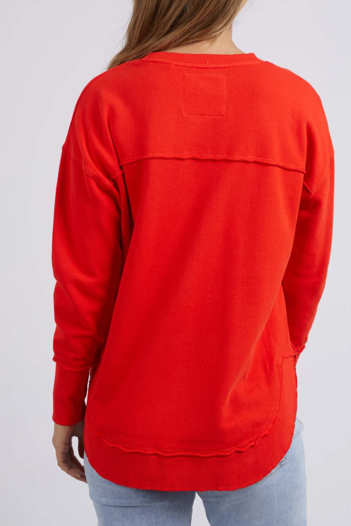 A person wears a bright red Foxwood Simplified Crew sweatshirt with a relaxed fit and rectangular stitch at the neckline. Made from 100% cotton with a curved hem, it's paired with light blue jeans against a plain gray background.