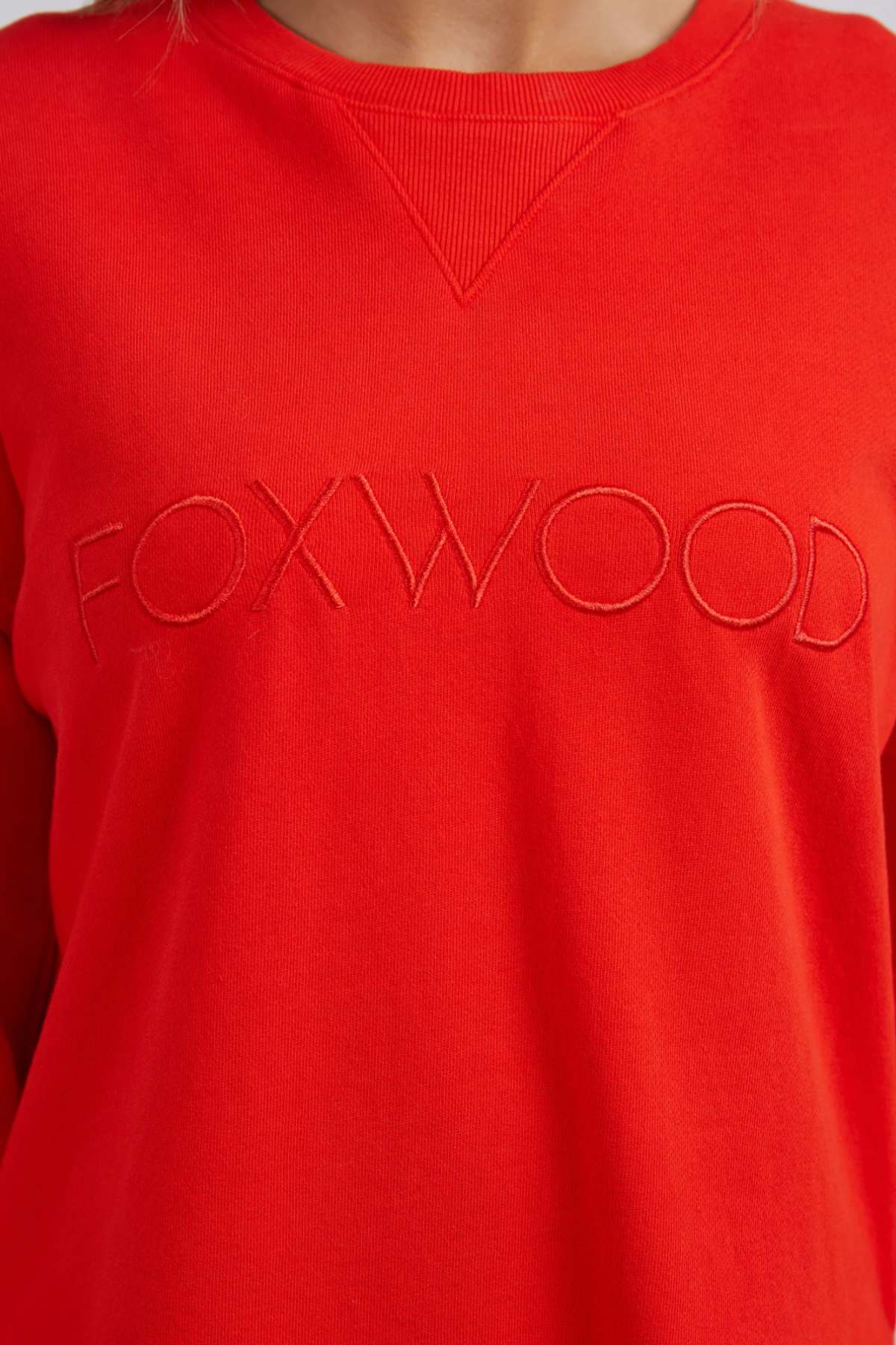 Close-up of a person wearing the relaxed fit Foxwood Simplified Crew, a bright red 100% cotton sweatshirt with "FOXWOOD" embroidered in red across the front and a triangular ribbed detail at the neckline.