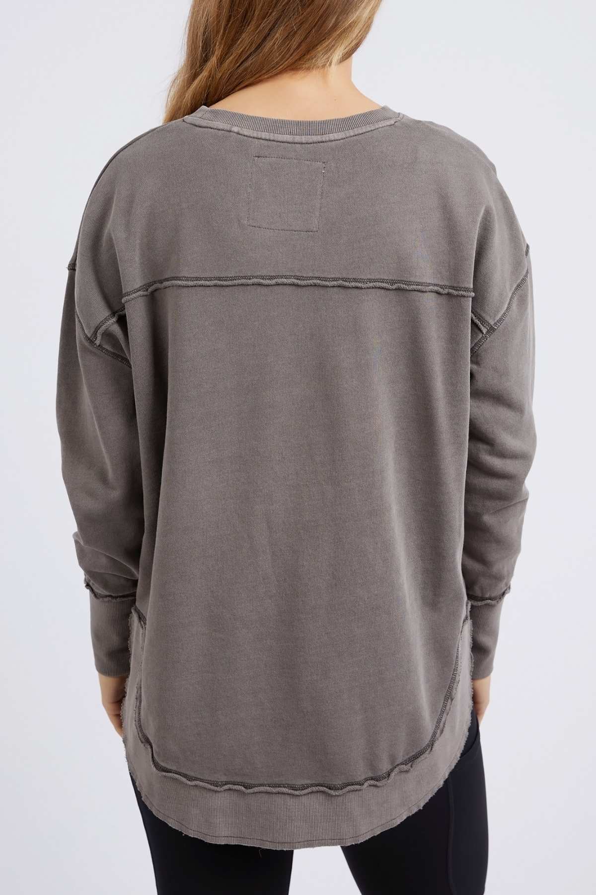 A person with long hair wears the Foxwood Simplified Crew, a relaxed-fit 100% cotton gray sweatshirt with exposed seams, paired with black leggings. They stand with their back to the camera against a plain white background.