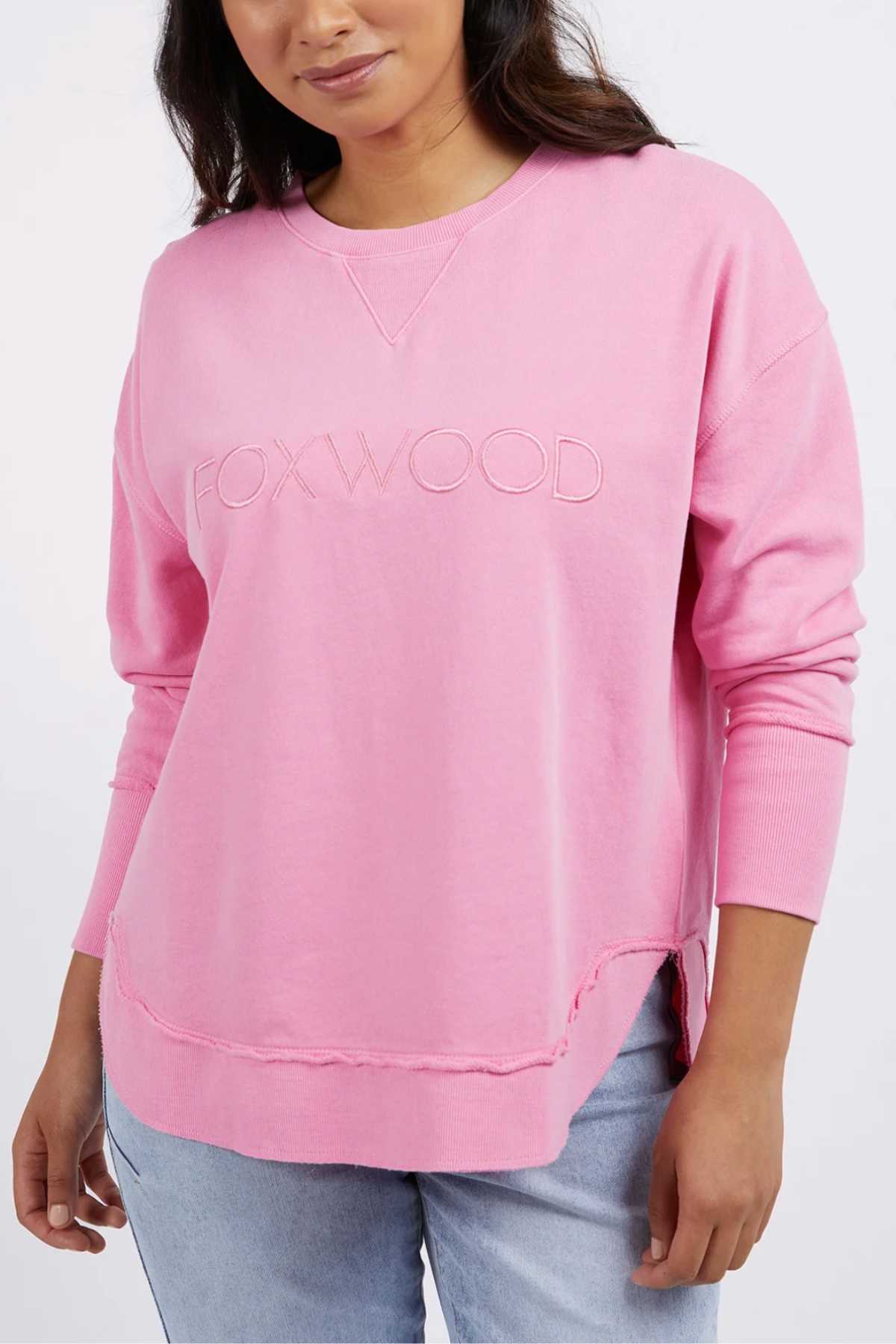 A woman wears the Foxwood Simplified Crew, a light pink 100% cotton sweatshirt with "FOXWOOD" embroidered on the front, standing with one hand relaxed at her side and paired with light blue jeans.