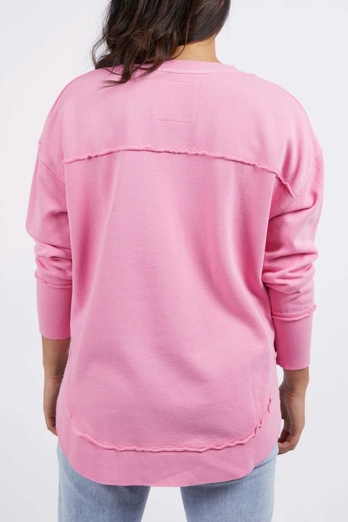 A person with long dark hair is seen from the back wearing the pink Foxwood Simplified Crew, featuring raw-edge seams, subtle embroidery, and a loose fit in 100% cotton. Light blue jeans complete the look against a plain background.