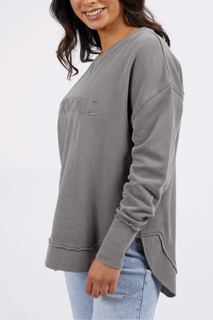 A person with dark hair wears a grey, long-sleeved Foxwood Simplified Crew sweatshirt adorned with a subtle embossed design and light blue jeans. They are standing and smiling, with their body slightly angled to the side, and their right hand resting by their hip.