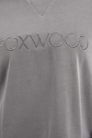 Close-up image of a grey 100% cotton crew neck with the word "FOXWOOD" embroidered in large, thin capital letters across the chest. The texture of the fabric and stitching details are visible, showcasing the fine craftsmanship of this Foxwood Simplified Crew.