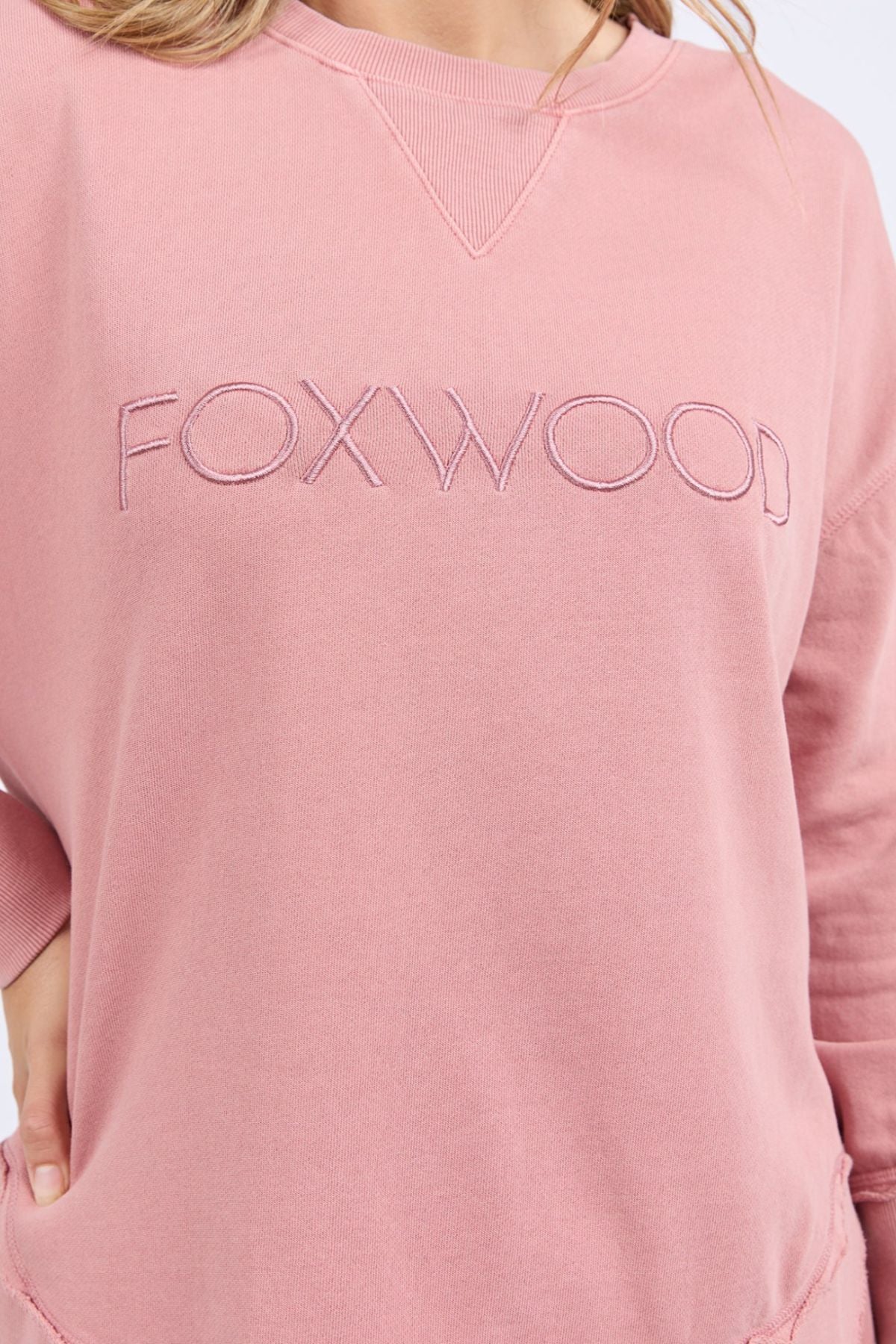 A person wears a relaxed fit, light pink Foxwood Simplified Crew, made from 100% cotton, with "FOXWOOD" embroidered in large capital letters across the chest. Only the torso and part of the arm are visible.