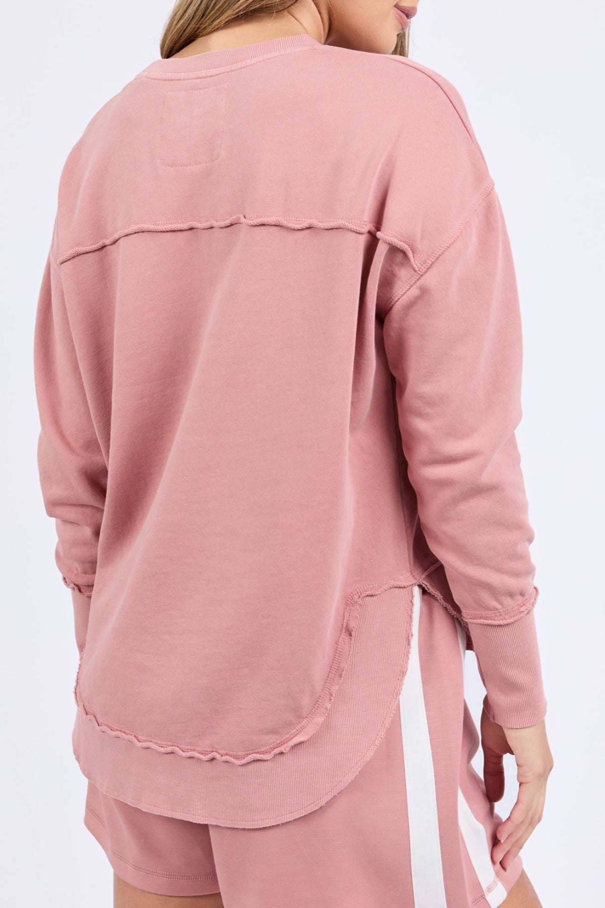 A person is shown from the back wearing the Foxwood Simplified Crew, a relaxed fit 100% cotton sweater in dusty pink with visible seams, paired with matching pink shorts with a white stripe, set against a plain background.