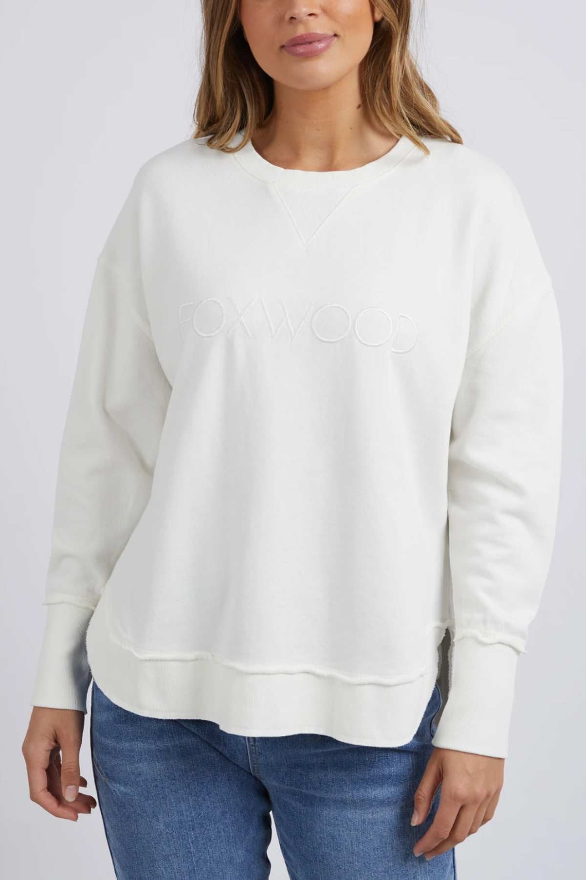 A person wearing a Foxwood Simplified Crew sweatshirt, featuring a light-colored design with "FOXWOOD" emblazoned on the front, stands against a neutral background. The outfit includes long sleeves and is paired with blue jeans. The person's hair is partially visible, resting on their shoulders.