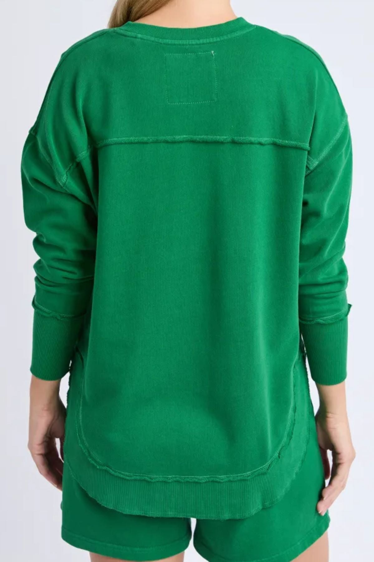 Back view of a woman wearing the Foxwood Simplified Crew, a relaxed-fit green cotton sweatshirt with exposed seam details, paired with matching green shorts.