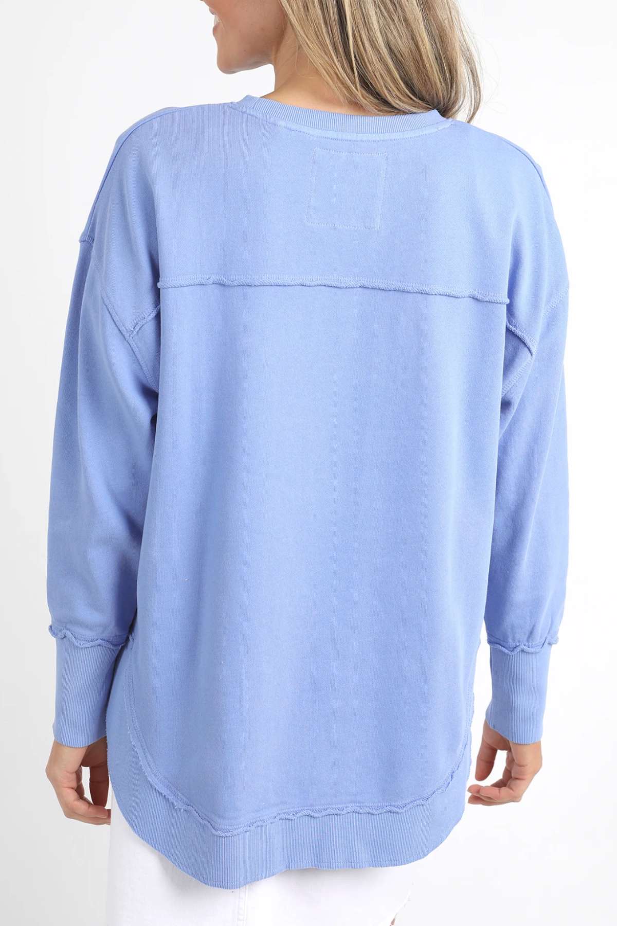 A person with long blonde hair is shown from the back, wearing a light blue, long-sleeved Foxwood Simplified Crew sweatshirt made from 100% cotton for comfort and breathability. They are also wearing white pants against a plain white background.