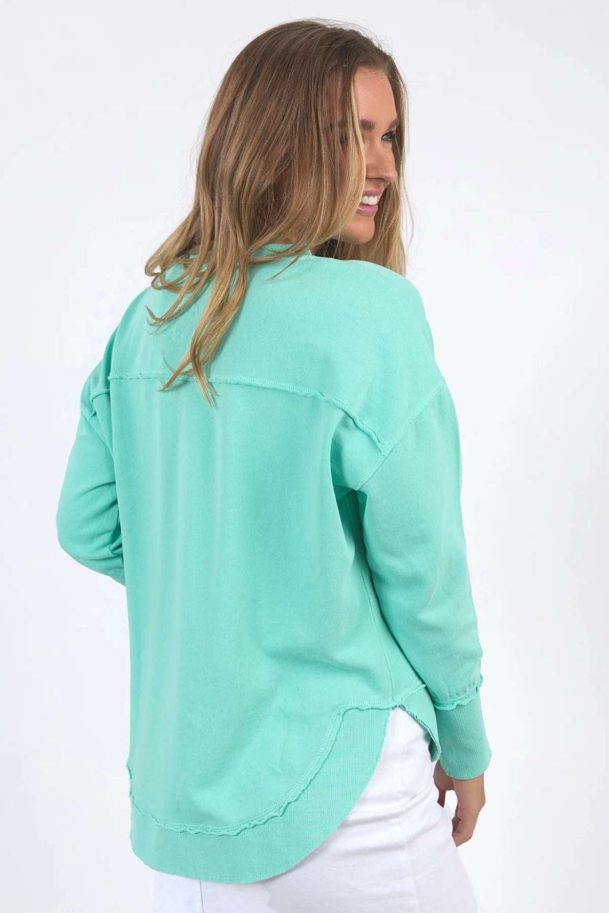 A woman with long, light brown hair smiles while looking over her shoulder. She is wearing a relaxed-fit Foxwood Simplified Crew sweater in mint green, crafted from 100% cotton, paired with white pants. The background is plain white.