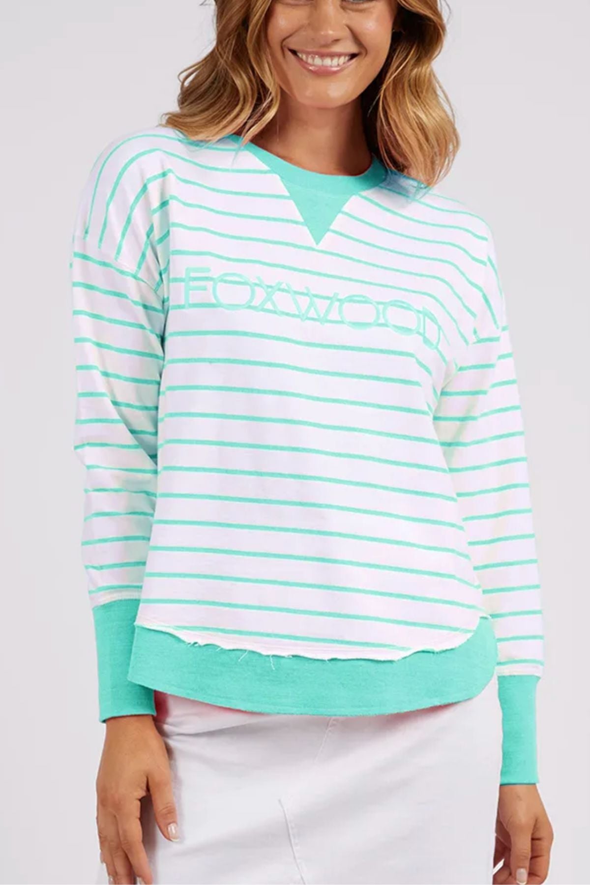 A woman is wearing a mint green and white striped "Foxwood Simplified Stripe Crew" sweater, which offers a relaxed fit and is crafted from 100% cotton. The word "Foxwood" is prominently displayed on the front, and the sweater is accented with a green collar and cuffs. She complements the look with a light-colored skirt, smiling against a plain background.