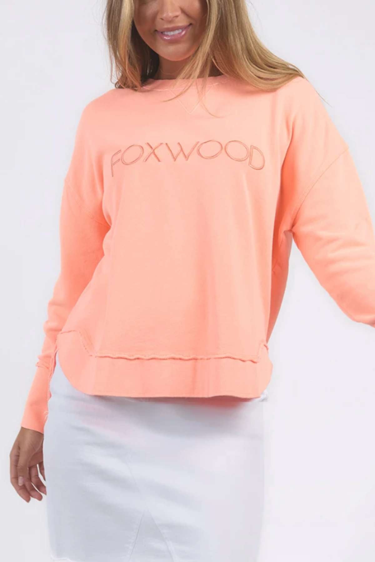 A person with long hair wears a light coral Foxwood Simplified Crew sweatshirt and a white skirt. The relaxed-fit, 100% cotton sweatshirt features a slightly frayed hem, adding to its casual look. The person is smiling and standing against a plain backdrop.