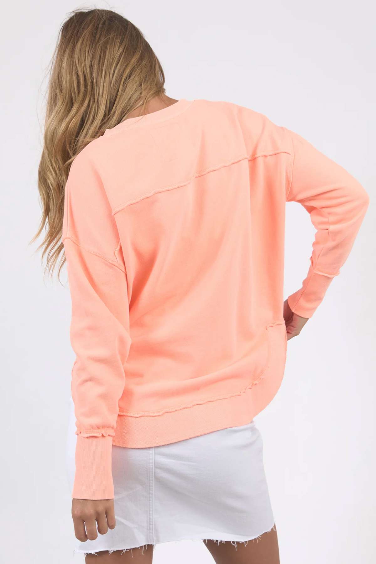 A person with long wavy hair stands with their back to the camera, wearing a relaxed fit, pastel peach Foxwood Simplified Crew and a white, frayed hem mini skirt. They have one hand on their hip and the other arm relaxed. The background is plain white.