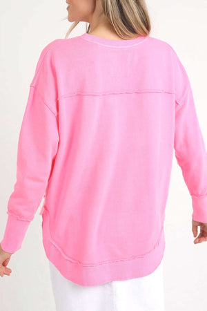 A person with long, light-colored hair wears the Foxwood Simplified Crew, a bright pink, relaxed-fit sweatshirt featuring a seam across the upper back and subtle distressing at the edges. Made from 100% cotton, it pairs perfectly with a white skirt that is partially visible as they face away from the camera.