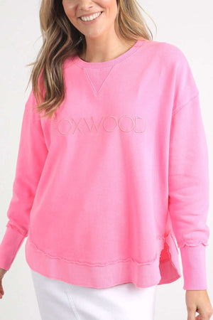 A smiling person with long, wavy hair is wearing a 100% cotton Foxwood Simplified Crew sweatshirt in bright pink, featuring "FOXWOOD" subtly embroidered on the front in matching pink thread. They are also dressed in a white skirt against a plain white background.