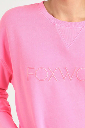 A person wears a vibrant pink Foxwood Simplified Crew sweatshirt, showcasing the partially visible "FOXWO" embroidery in light pink on the front. The relaxed fit of this 100% cotton garment is complemented by its ribbed collar and a distinctive triangular patch detail underneath. The background is plain white.