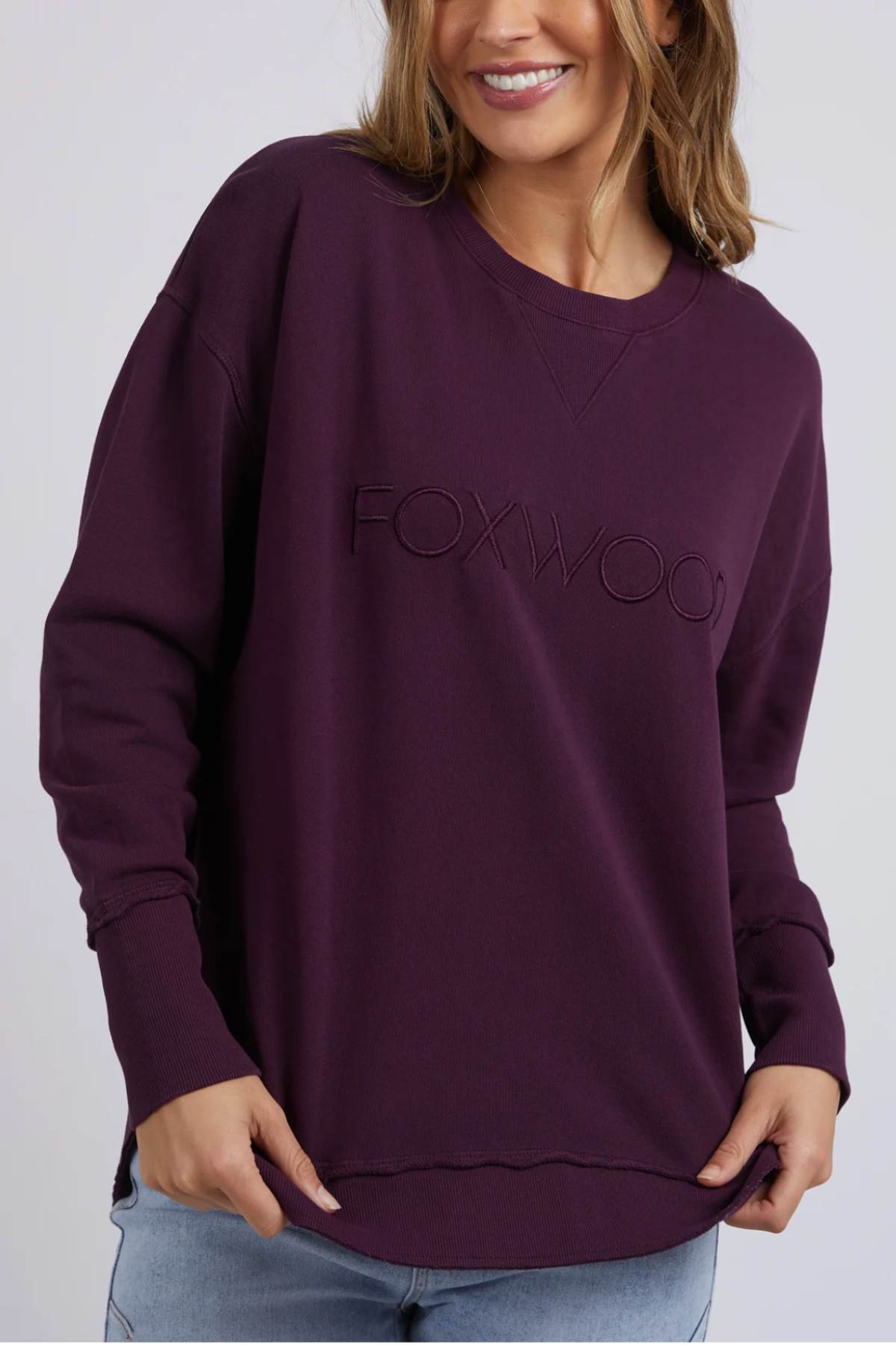A woman wearing a relaxed fit, 100% cotton Foxwood Simplified Crew sweatshirt in dark purple with "FOXWOOD" embroidered on the front, paired with light blue jeans, smiles with her hands near her hips.