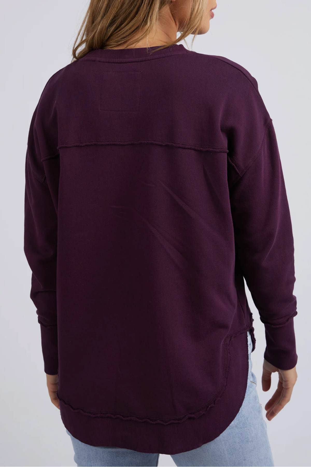 A person is seen from the back wearing a long-sleeved, dark purple Foxwood Simplified Crew sweatshirt with exposed seams and light blue jeans. The relaxed, 100% cotton fit adds comfort to this casual look.