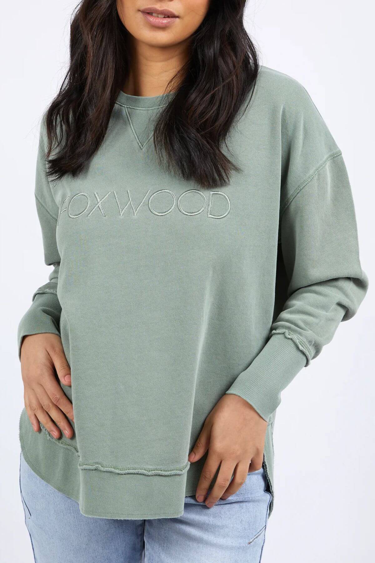 A woman with wavy dark hair is wearing a relaxed-fit, light green sweatshirt emblazoned with "FOXWOOD" on the front. This 100% cotton Foxwood Simplified Crew has one of her hands on her hip and the other resting on her thigh. She pairs it with light blue jeans against a plain white background.