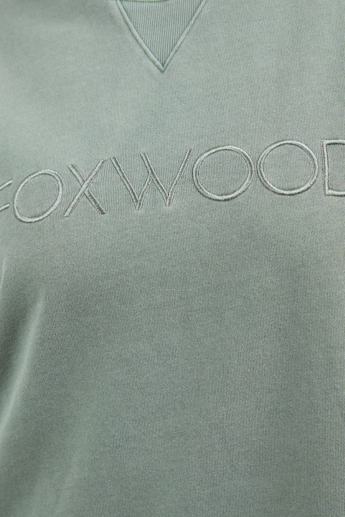 A close-up image of a sage green sweatshirt displaying the text "FOXWOOD" embroidered across the chest in matching green thread. The neckline features a ribbed V-shaped detail. This Foxwood Simplified Crew is made from 100% cotton and offers a relaxed fit for ultimate comfort.