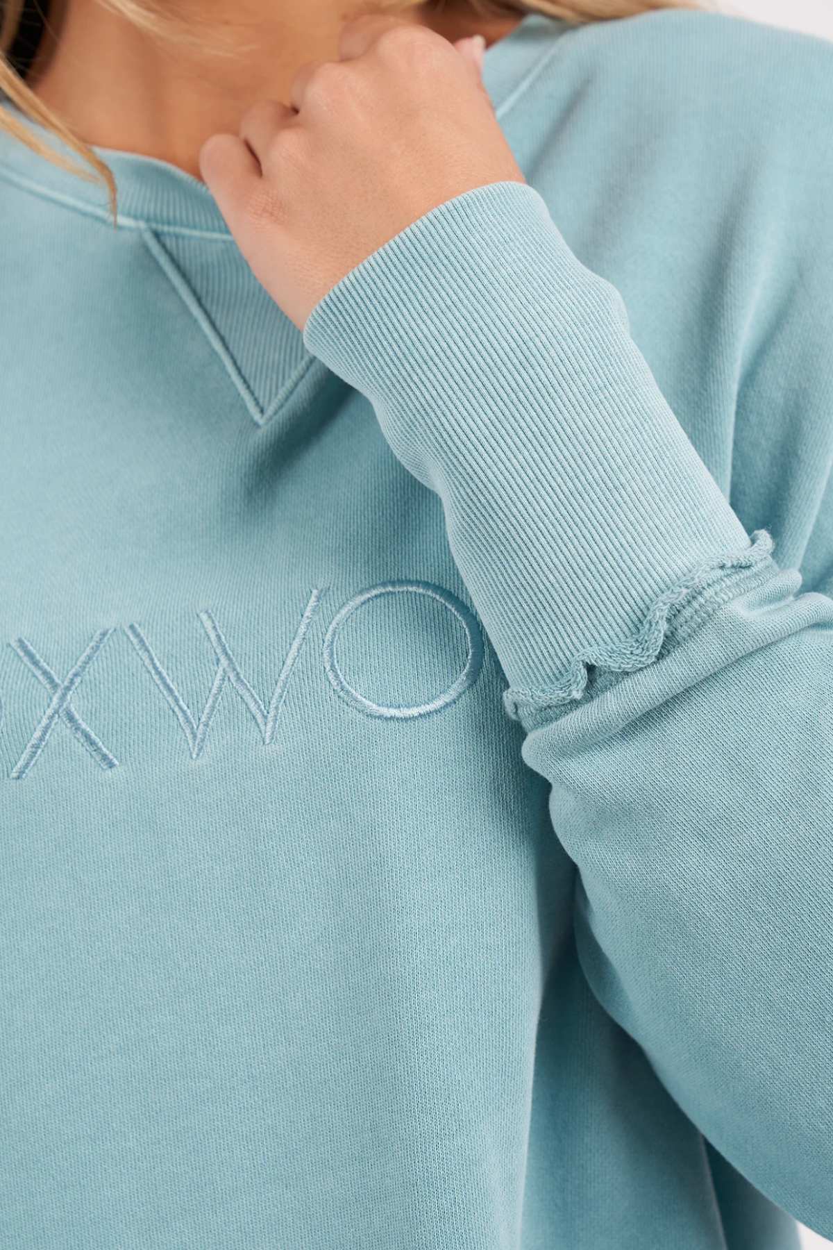 Close-up of a person wearing the Foxwood Simplified Crew sweatshirt in light blue with ribbed cuffs. The embroidered partial text reads "XWO." The person's hand is slightly raised near their chin, highlighting the fabric's texture.