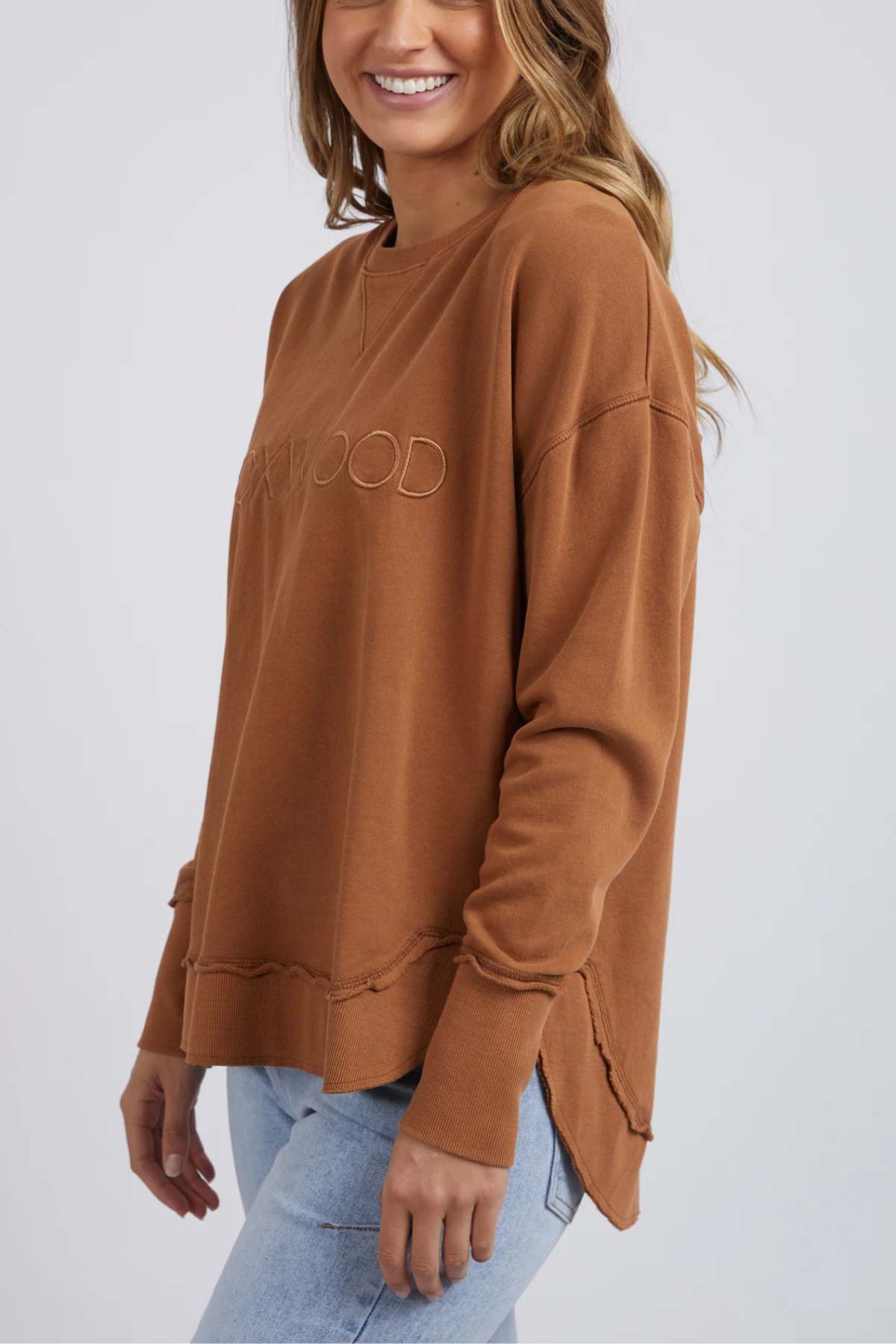 A woman smiles in a loose brown Foxwood Simplified Crew, featuring subtle embroidery, paired with light blue jeans against a plain light background.