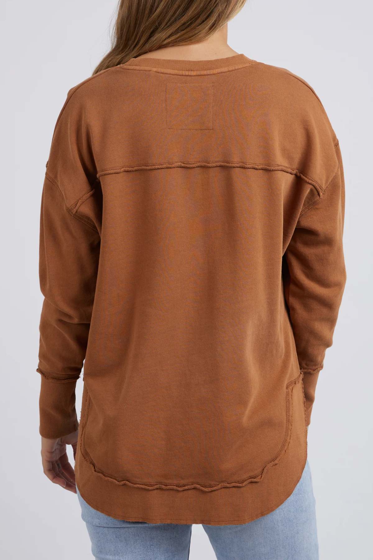 A person with long brown hair, shown from the back, wears a brown Foxwood Simplified Crew made of 100% cotton with visible seams, paired with light blue jeans.