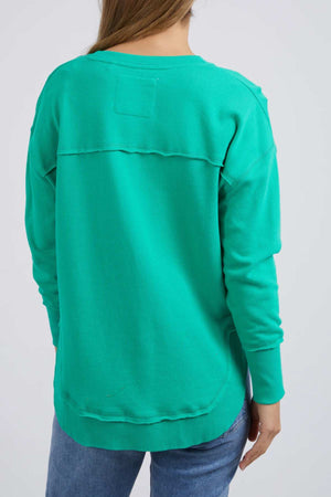 A person with straight brown hair is shown from the back, wearing a bright green Foxwood Simplified Metallic Crew sweatshirt with a visible upper-back seam, paired with blue jeans.