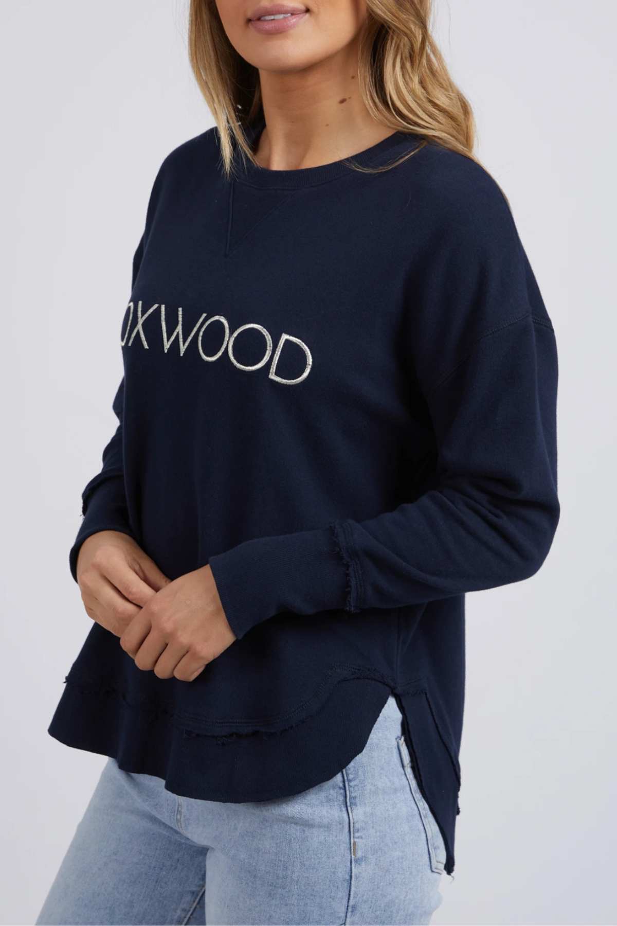 A woman wears the Foxwood Simplified Metallic Crew in navy cotton with "FOXWOOD" white chest embroidery, paired with light blue jeans. Her hair is loose and she stands against a plain light background.