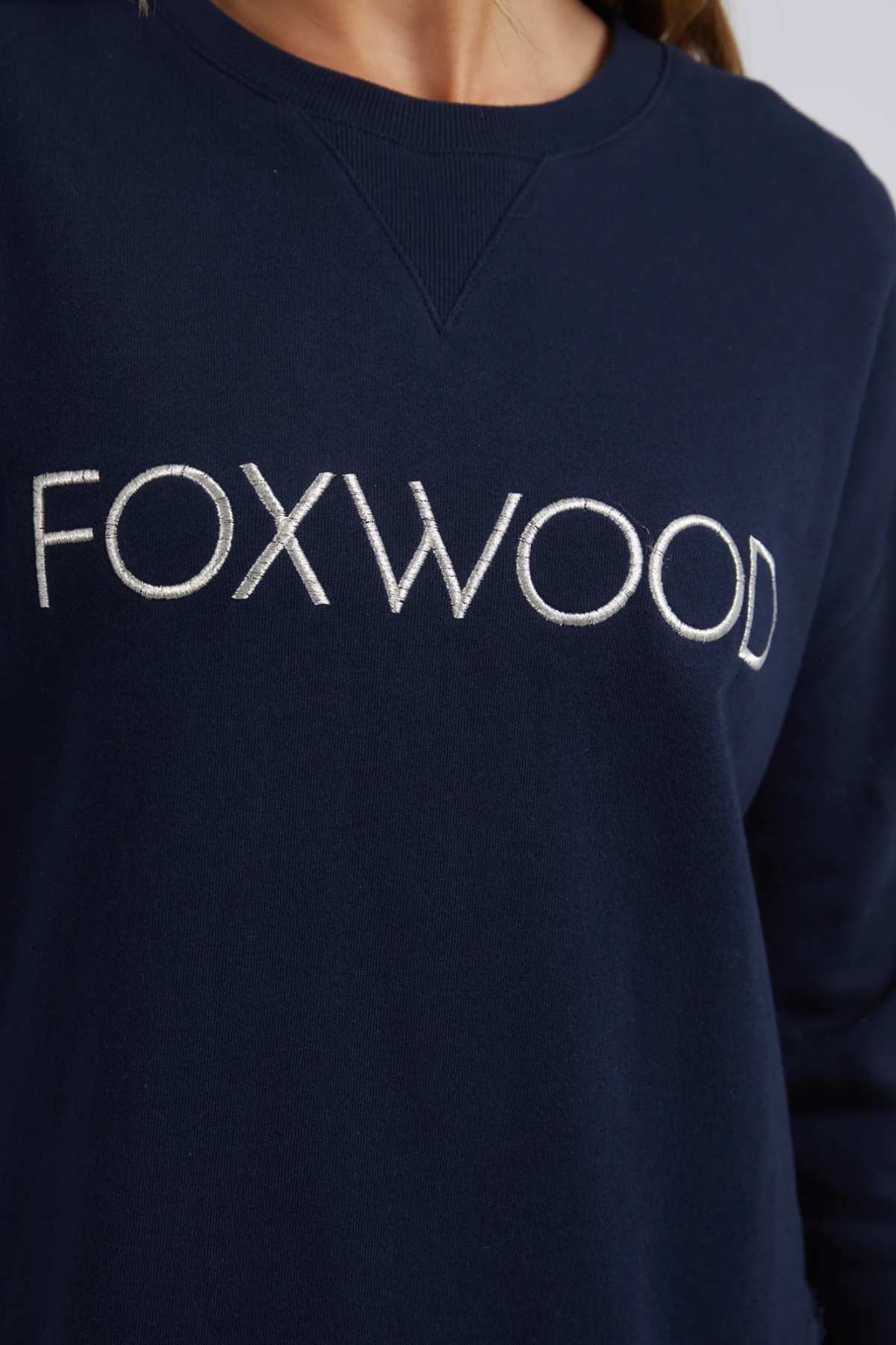 A person wearing a dark blue Foxwood Simplified Metallic Crew, made of cotton, featuring "FOXWOOD" embroidered in white on the chest.