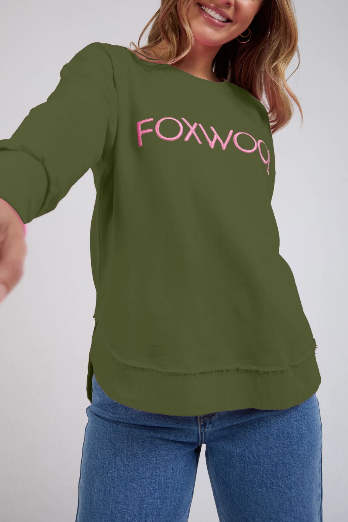 A smiling woman with wavy hair wears a green Foxwood Simplified Metallic Crew with pink Foxwood embroidery and blue jeans, reaching one arm toward the camera.
