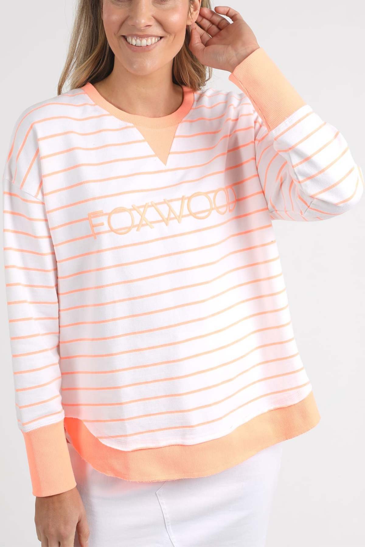 Wearing the Foxwood Simplified Stripe Crew, a white and peach-striped sweater adorned with intricate Foxwood embroidery, a person gently touches their hair while smiling. This classic crew neck sweater is crafted from 100% cotton and is perfectly paired with white bottoms against a light-colored background.
