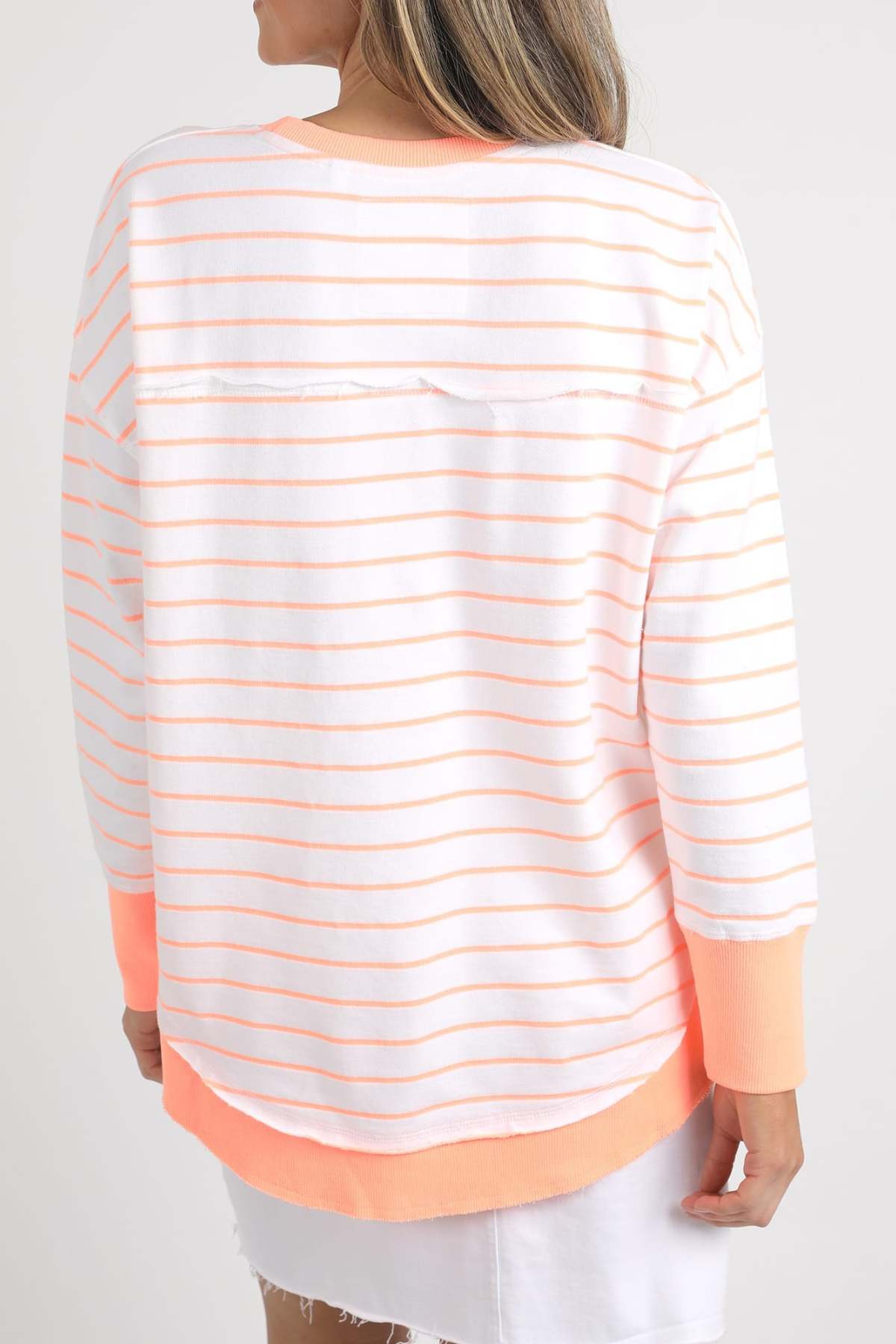 A person is wearing the Foxwood Simplified Stripe Crew, a 100% cotton white sweater adorned with thin orange horizontal stripes and subtle Foxwood embroidery. This slightly loose-fit, crew neck design includes pink-orange cuffs and hem. They are facing away while pairing the sweater with a white skirt.