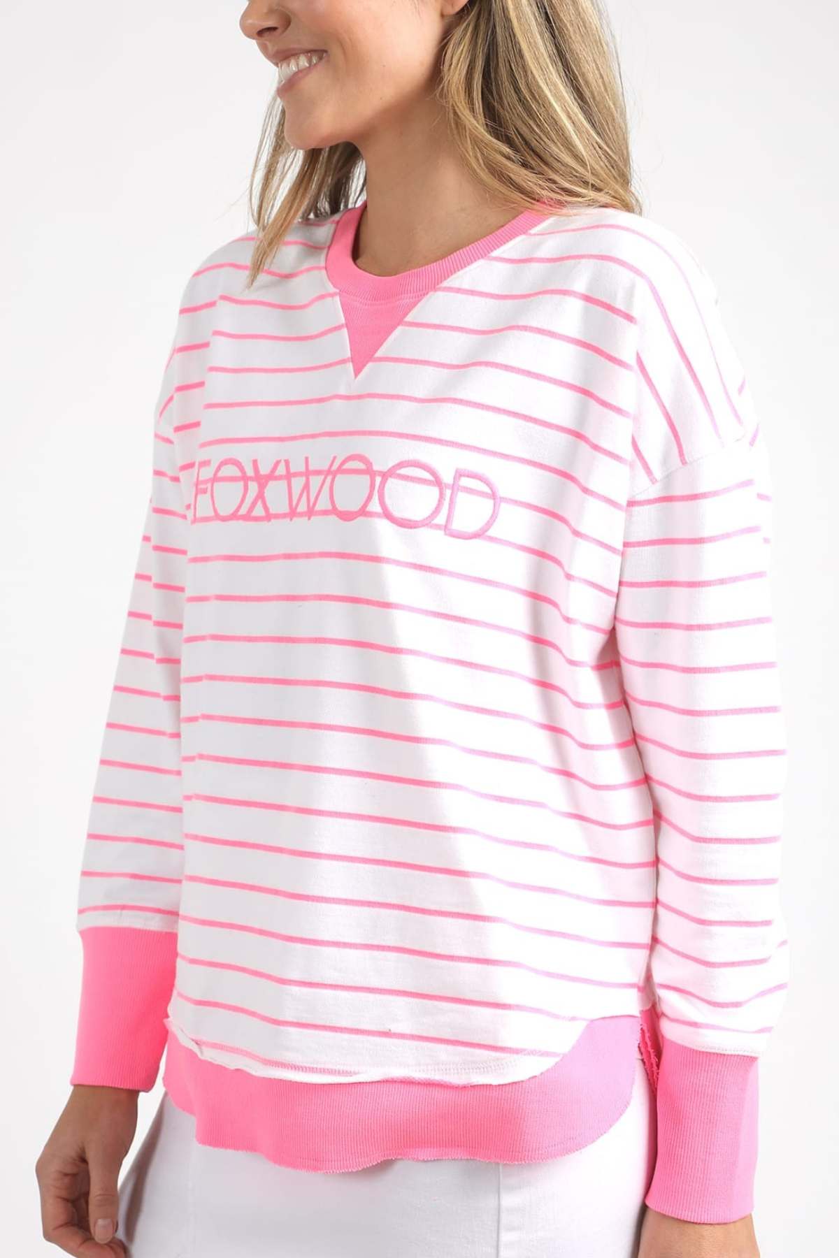 The individual is donning the Foxwood Simplified Stripe Crew, a relaxed fit sweatshirt made from 100% cotton, showcasing "FOXWOOD" on the front. Its white and pink striped pattern features pink cuffs and a neckline, embodying the signature Foxwood Crew style. They are smiling warmly against a plain backdrop.