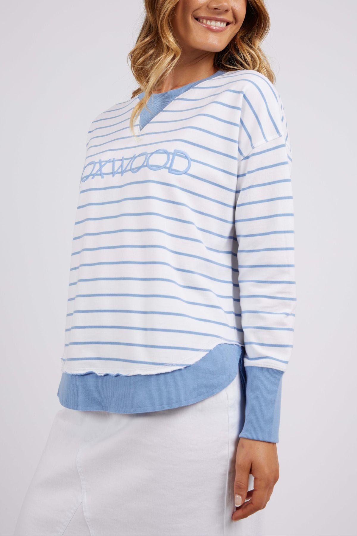 A person is wearing the Foxwood Simplified Stripe Crew, which showcases white and light blue yarn-dyed stripes, along with "OXWOOD" embroidered on the front. The sweater's sophisticated blue cuffs and hem are beautifully matched with a white skirt. With long hair cascading down, they are beaming joyfully.