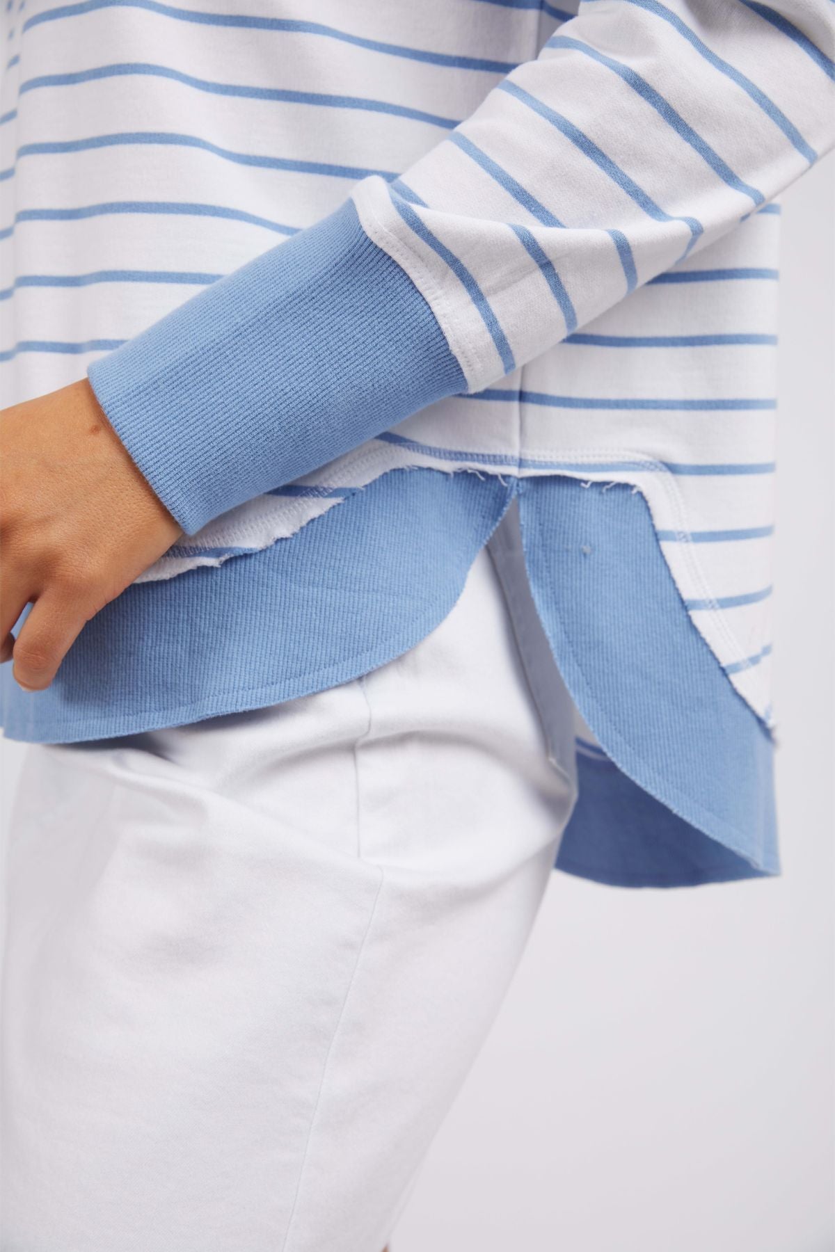 Dressed in the Foxwood Simplified Stripe Crew, which showcases blue and white yarn-dyed stripes along with light blue cuffs and a distinctive layered hem, an individual is posed slightly to the side. Complemented by sharp white pants, this ensemble perfectly embodies the signature style of Foxwood Clothing.