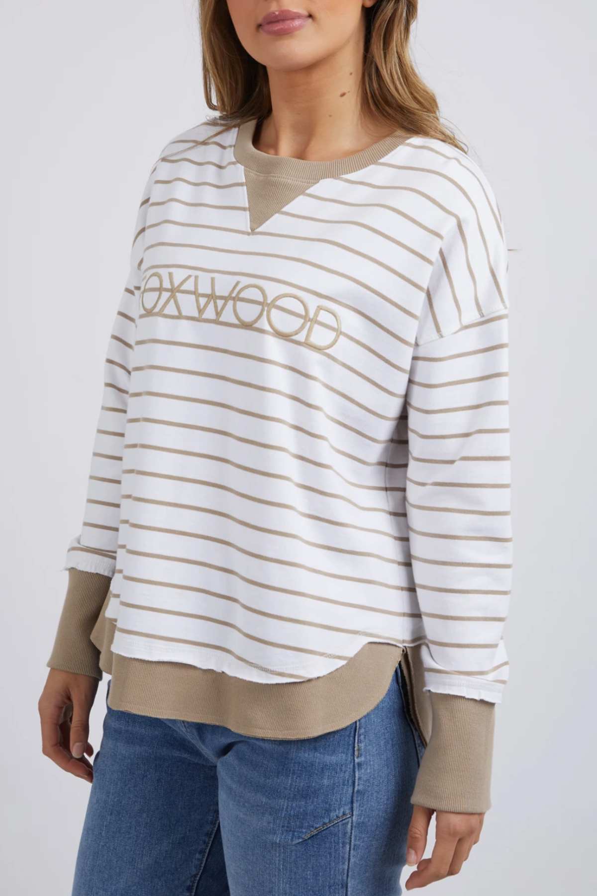 A person is wearing the Foxwood Simplified Stripe Crew in white and tan, showcasing "FOXWOOD" on the front. This relaxed-fit, 100% cotton sweatshirt with tan cuffs and collar is paired with blue jeans against a plain white background.