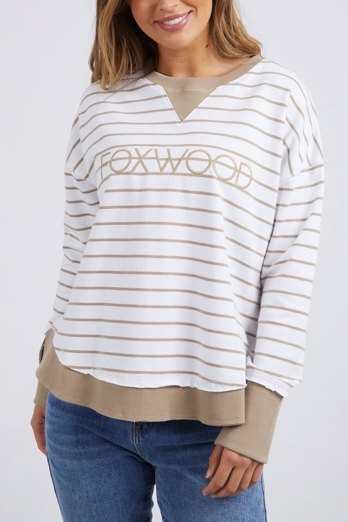 The woman is dressed in the Foxwood Simplified Stripe Crew, showcasing subtle white and beige yarn dye stripes with "FOXWOOD" tastefully displayed on the front. The beige cuffs and hem of the shirt harmonize beautifully with her blue denim jeans, embodying Foxwood Clothing's casual sophistication.