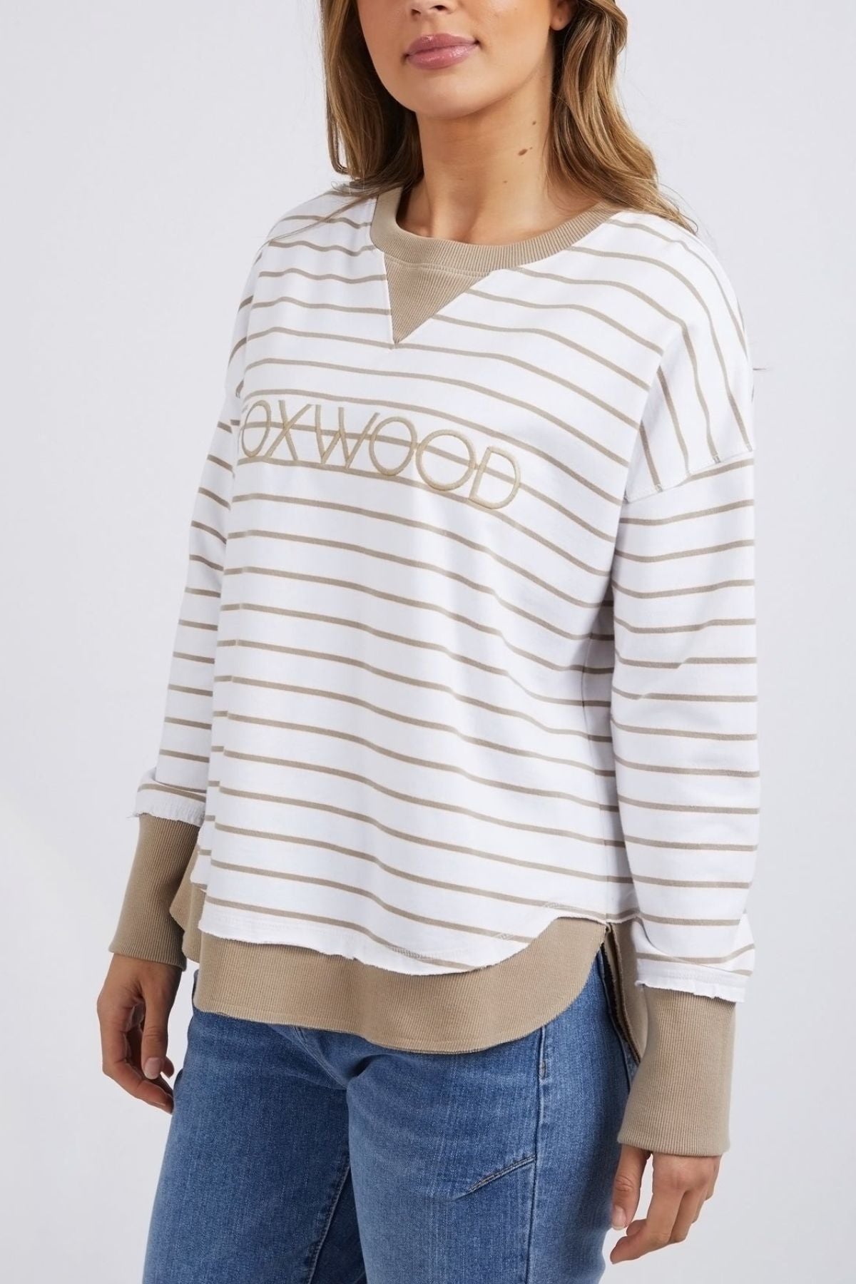 A person is dressed in the Foxwood Simplified Stripe Crew, which showcases white and beige yarn-dyed stripes and prominently displays "OXWOOD" on its front. This round neck sweater comes with beige cuffs and is stylishly paired with blue jeans, creating a striking contrast against a simple backdrop.