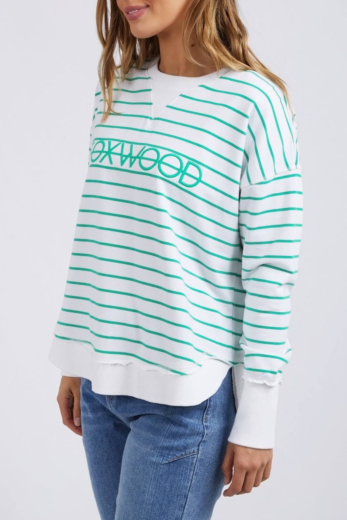 A person is wearing a white and green striped "Foxwood Simplified Stripe Crew" sweater, crafted from 100% cotton for a relaxed fit, paired with blue jeans. The crew style complements their wavy brown hair as they stand against a plain white background.